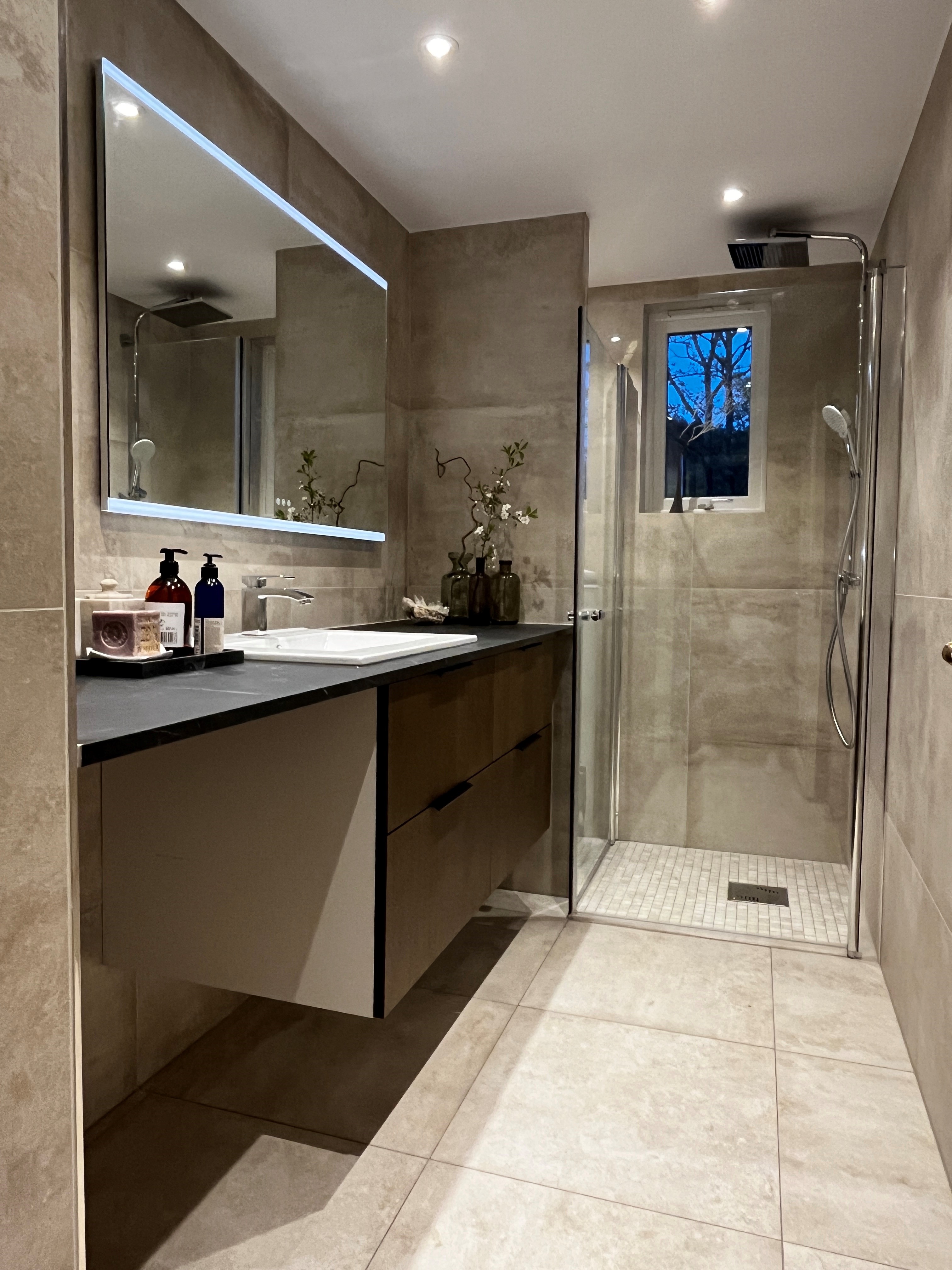 Modern bathroom with light gray tiled walls and floor, an illuminated mirror, a white sink with a chrome faucet on a dark countertop, and a floating vanity with drawers. The room features a glass-walled shower area and various bathroom accessories on the countertop. A small window is visible in the shower area.