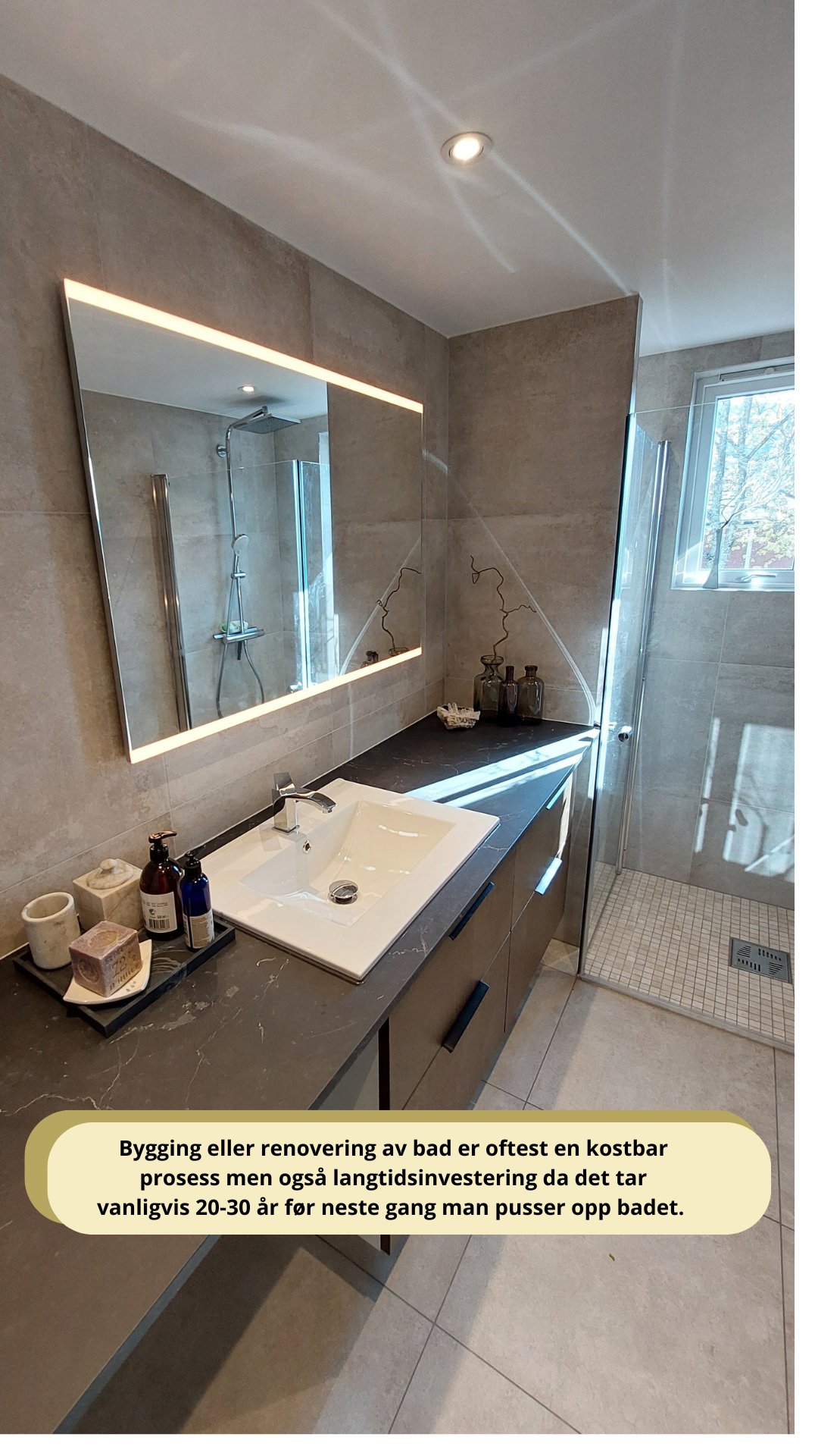 Modern bathroom with a large illuminated mirror, light gray tiled walls, a white rectangular sink with a chrome faucet on a dark countertop, and a glass-walled shower area. Various bathroom accessories are neatly arranged on the countertop. Natural light is streaming in through a window near the shower.